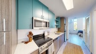 This efficiently designed one-bedroom, one-bathroom unit makes excellent use of its compact footprint, offering a well-organized and modern living environment across a single floor. The galley-style kitchen is a standout feature, showcasing bold teal upper cabinetry paired with warm wood-toned lower cabinets and crisp white quartz countertops. A full suite of stainless steel appliances — including an electric range, over-the-range microwave, dishwasher, and refrigerator — lines the kitchen, while a skylight overhead draws in natural light to brighten the space. The bedroom occupies roughly half the unit's total area and is highlighted by a bank of tall, floor-to-ceiling windows that flood the room with natural light and offer views of surrounding greenery. Light wood-tone flooring runs throughout, contributing to a clean, airy aesthetic. A dedicated open closet nook with a wall-mounted clothing rail provides practical storage within the bedroom area. The bathroom is cleanly appointed with a tub-shower combination, a wood-grain vanity with stone-look countertop, and neutral tile flooring. The unit's modern design, cohesive color palette, and efficient layout make it a well-suited space for comfortable single-occupant living.
