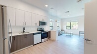 Located at 345 Lincoln Ave in Amherst, MA, this single-floor unit offers a thoughtfully designed layout that maximizes its compact footprint. The open-plan kitchen and living area form the heart of the space, finished throughout with light hardwood-style flooring that enhances the bright, airy atmosphere.

The kitchen is outfitted with a two-tone cabinet scheme—white upper cabinets contrasting with dark wood-grain lower cabinets—accented by matte black hardware. Stainless steel appliances include a full-size refrigerator, electric range, microwave, and dishwasher, complemented by white quartz countertops and a single-basin sink with a sleek black faucet. A peninsula with seating provides a natural transition into the living area.

The living area receives ample natural light through large windows framing views of surrounding greenery, creating a calm, inviting atmosphere. Recessed and pendant lighting provide well-distributed illumination throughout.

A hallway connects to the bathroom, which features a wood-grain vanity with white countertop, a walk-in shower, and coordinating light hardwood-style flooring. Clean white walls and minimalist finishes are consistent throughout, giving the entire space a cohesive, contemporary character.