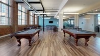 This expansive game room blends industrial character with modern amenities, offering a well-appointed recreational retreat. The space is defined by soaring ceilings with exposed ductwork and structural columns, original brick walls, and oversized factory-style windows that flood the interior with natural light and frame views of the surrounding cityscape. Warm hardwood flooring runs throughout, complementing the raw architectural elements.

The room is generously outfitted for entertainment, featuring multiple full-size billiard tables with slate-blue felt, a regulation ping-pong table, a foosball table, and a shuffleboard table — providing a diverse range of gaming options. High-top bar tables and stools are positioned throughout, offering casual seating near the windows and along accent walls painted in a muted teal tone.

A floor-to-ceiling glass partition wall borders one side of the space, offering visual connectivity to an adjacent fitness or lounge area while maintaining acoustic separation. An arched doorway provides a refined architectural detail, leading to a quieter nook with bar seating and outdoor views. The overall ambiance is dynamic yet inviting, merging historic building character with contemporary recreational design.