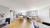 This well-appointed single-floor communal space offers a thoughtfully designed open-concept layout that seamlessly integrates a kitchen, dining, and lounge areas. The kitchen is outfitted with modern grey cabinetry, white upper cabinets, stainless steel appliances including a dishwasher and microwave, a double sink, and a striking mosaic tile backsplash — all anchored by clean white countertops. A window above the kitchen counter fills the workspace with natural light and outdoor views of surrounding greenery and buildings.

The main floor area features warm light wood flooring throughout, lending a cohesive and inviting feel to the space. Multiple seating and dining configurations are present, including a round white table with modern chairs and additional folding table setups, making the space highly adaptable for group gatherings or individual use. A well-stocked bookshelf lines one wall, adding a resourceful and community-oriented touch.

The lounge area is anchored by a built-in wood-paneled entertainment unit housing a wall-mounted television and electric fireplace, complemented by vibrant teal accent chairs and a grey sofa. Large windows along the perimeter flood the interior with natural light while offering open views of the surrounding neighbourhood, contributing to the space's bright and welcoming ambiance.