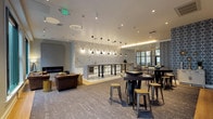 This single-floor amenity lounge is situated within a landmark arcade building at 161 West Wisconsin Avenue in Milwaukee, identifiable by its stunning glass-domed rotunda, ornate ironwork balconies, and grand spiral staircase visible through the lounge's glass entry doors. The interior blends contemporary design with the building's storied architectural character.

The open-plan layout is anchored by a long, white-topped wet bar illuminated by a row of sleek black pendant lights, with gold wire barstools lining its face and decorative stone-textured wall cladding behind. Adjacent to the bar, a social seating area features high-top black tables and industrial metal stools, ideal for casual gathering.

A lounge nook offers plush, gold-framed chevron-upholstered chairs and a tufted ottoman arranged around a textured accent wall, creating an intimate setting. Nearby, a tufted leather Chesterfield sofa faces oversized black-framed windows that frame direct city skyline views, flooding the space with natural light.

A linear fireplace anchors a more relaxed corner with leather club seating. Patterned carpeting, warm wood flooring accents, and layered lighting throughout contribute to the lounge's polished yet welcoming ambiance.