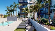 Located at 1725 W Katella Ave in Orange, California, this thoughtfully designed outdoor amenity space offers a resort-inspired experience within a modern residential community. The space is anchored by a striking double-sided brick fireplace that serves as both a functional centerpiece and a architectural divider, creating two distinct lounge areas furnished with bold yellow curved sofas and accent chairs, paired with natural wood side tables atop artificial turf. The warm, inviting atmosphere of the fireplace lounge transitions seamlessly into an expansive poolside area, where rows of sleek charcoal chaise lounges are arranged along a sun-drenched deck. Towering palm trees frame the pool area, lending a tropical ambiance while providing gentle shade throughout the day. The surrounding landscaping features drought-tolerant plants and lush greenery that complement the clean, contemporary architecture of the multi-story building rising above. Sculptural white pendant elements add artistic flair to the outdoor environment. The overall design balances comfort and style, creating an engaging outdoor retreat ideal for relaxation and social gatherings, all under the abundant natural light characteristic of Southern California.