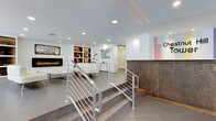 This contemporary commercial space makes a strong first impression with its thoughtfully designed lobby and reception area. Upon entry, visitors are greeted by a polished wooden reception desk accented by a bold, colorful graphic logo, setting a professional yet inviting tone. The space transitions seamlessly into a lounge area, elevated on a platform and accessible via wide steps framed by sleek metal railings with horizontal cable detailing.

The lounge area is a standout feature, anchored by a modern electric fireplace built into a crisp white wall, casting a warm ambient glow. Flanking the fireplace are open built-in shelving niches displaying decorative objects, adding visual interest without cluttering the clean aesthetic. A low-profile white sofa and minimalist coffee table complete the seating arrangement, offering a comfortable area for guests.

The floors throughout are finished in large-format neutral tiles, contributing to the spacious, open feel of the interior. Recessed ceiling lighting provides even, bright illumination across the entire space. The white walls are punctuated by carefully placed artwork, adding color and personality. The overall design is modern and minimalist, balancing functionality with a welcoming ambiance suited to a professional commercial environment.