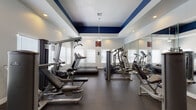 This fitness center offers a comprehensive and thoughtfully designed workout environment suited for a variety of exercise routines. The space is defined by its striking tray ceiling, accented with a bold navy blue border that contrasts cleanly against the white walls, lending the room a polished and energetic aesthetic. A ceiling fan centered in the tray provides air circulation, while recessed lighting ensures the space is well-lit throughout.

Large windows with white blinds line multiple walls, allowing generous natural light to fill the room and create an open, airy atmosphere. Full-length wall mirrors on one side visually expand the space and allow users to monitor their form during workouts.

The equipment selection is extensive and well-maintained. Cardio machines include multiple treadmills and Precor elliptical trainers, providing ample options for aerobic exercise. Strength training is supported by a cable crossover machine, a multi-function weight machine, and a dedicated free weights rack stocked with dumbbells and yoga mats for stretching and floor exercises. Durable rubber flooring throughout the space provides traction and impact absorption.

The fitness center is accessed through elegant French doors and includes posted fitness center rules, a sanitizing station, and clearly marked emergency signage, reflecting attention to both safety and user experience.