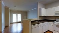 This single-floor residence offers a thoughtfully designed open-concept layout that seamlessly connects the kitchen and living areas. The kitchen is well-appointed with crisp white cabinetry providing ample storage, dark countertops, a double-basin sink, a dishwasher, and a peninsula that serves both as a functional workspace and a natural divider between the cooking and living zones. Crown molding along the ceiling adds a refined architectural touch throughout the space. The living area features warm hardwood flooring that extends throughout, enhancing the sense of openness and continuity. A pair of classic French doors floods the space with natural light, creating a bright and welcoming ambiance while offering a connection to the outdoors. The soft, neutral wall tones complement the white trim and cabinetry, resulting in a clean, cohesive aesthetic. The open floor plan allows for flexible furniture arrangements, making the living space both practical and inviting. Overall, this residence combines functional design with understated elegance, ideal for comfortable everyday living.