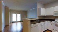 This single-floor residence offers a thoughtfully designed open-concept layout that seamlessly connects the kitchen and living areas. The kitchen is well-appointed with crisp white cabinetry providing ample storage, dark countertops, a double-basin sink, a dishwasher, and a peninsula that serves both as a functional workspace and a natural divider between the cooking and living zones. Crown molding along the ceiling adds a refined architectural touch throughout the space. The living area features warm hardwood flooring that extends throughout, enhancing the sense of openness and continuity. A pair of classic French doors floods the space with natural light, creating a bright and welcoming ambiance while offering a connection to the outdoors. The soft, neutral wall tones complement the white trim and cabinetry, resulting in a clean, cohesive aesthetic. The open floor plan allows for flexible furniture arrangements, making the living space both practical and inviting. Overall, this residence combines functional design with understated elegance, ideal for comfortable everyday living.
