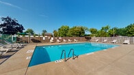 This outdoor amenity space serves as the central recreational hub of a multi-story residential building complex. The focal point is a generously sized rectangular swimming pool with clearly marked depth indicators, accessible via sturdy stainless steel entry rails. The pool's vivid turquoise water is complemented by a wide surrounding concrete deck, offering ample room for sunbathing and leisure activities.

Lining the perimeter of the deck are numerous lounge chairs, providing abundant seating for residents to relax in the open air. A shaded picnic area with a table and umbrella offers a comfortable space for outdoor dining and socializing. A small utility shed is positioned along the fence line, supporting pool maintenance needs.

The pool area is enclosed by a wooden privacy fence on multiple sides and a chain-link fence section that faces the adjacent parking lot, with mature trees visible beyond the boundary, adding a natural backdrop. The multi-story building rises prominently along one side, with its rows of balconies and large windows overlooking the pool area. The overall setting is open, sun-drenched, and practical, offering residents a well-equipped outdoor retreat well-suited for warm-weather enjoyment.