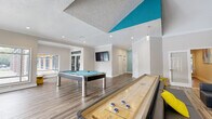 This single-floor amenity space is designed for recreation and social gathering, combining distinct activity zones within an open, well-lit layout. The interior is unified by wood-look plank flooring, crisp white walls, and a striking teal color scheme applied to ceiling panels, columns, and accent walls, lending the space a modern, energetic character throughout.

The game room occupies one section of the space, centered around a contemporary pool table with chrome legs and teal felt, complemented by a shuffleboard table. Recessed lighting and a wall-mounted flat-screen television enhance the entertainment-focused atmosphere. A vaulted ceiling with a bold teal triangular accent element rises above the adjacent lounge area, which features low-profile charcoal modular seating, yellow accent stools, and an additional wall-mounted television, creating a comfortable setting for group gatherings.

A sunlit dining area is enclosed by floor-to-ceiling windows on multiple sides, offering unobstructed views of an outdoor pool and courtyard. Tall bistro-style tables paired with swivel bar stools furnish this bright zone, which benefits from abundant natural light. A separate glass-enclosed fitness room is also accessible from within the space, rounding out the range of amenities available.