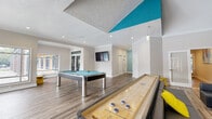 This single-floor amenity space is designed for recreation and social gathering, combining distinct activity zones within an open, well-lit layout. The interior is unified by wood-look plank flooring, crisp white walls, and a striking teal color scheme applied to ceiling panels, columns, and accent walls, lending the space a modern, energetic character throughout.

The game room occupies one section of the space, centered around a contemporary pool table with chrome legs and teal felt, complemented by a shuffleboard table. Recessed lighting and a wall-mounted flat-screen television enhance the entertainment-focused atmosphere. A vaulted ceiling with a bold teal triangular accent element rises above the adjacent lounge area, which features low-profile charcoal modular seating, yellow accent stools, and an additional wall-mounted television, creating a comfortable setting for group gatherings.

A sunlit dining area is enclosed by floor-to-ceiling windows on multiple sides, offering unobstructed views of an outdoor pool and courtyard. Tall bistro-style tables paired with swivel bar stools furnish this bright zone, which benefits from abundant natural light. A separate glass-enclosed fitness room is also accessible from within the space, rounding out the range of amenities available.