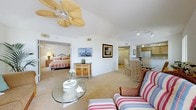 This single-floor residence offers a thoughtfully designed layout with a warm, coastal ambiance throughout. The open-concept living and dining area serves as the heart of the home, featuring soft carpet flooring, a decorative ceiling fan, and a wall of sliding glass doors that flood the space with natural light and provide direct access to an outdoor balcony with pleasant exterior views. Tropical-themed artwork and light, neutral tones reinforce the relaxed coastal character of the space.

The dining area connects seamlessly to the kitchen, which is separated by a pass-through counter. The kitchen is outfitted with light wood cabinetry, white appliances including a range, microwave, and refrigerator, and rich hardwood flooring that contrasts nicely with the surrounding carpeted areas. Track lighting overhead ensures ample illumination throughout.

The primary bedroom is generously proportioned, featuring a tray ceiling, plush carpeting, a large closet, and an en-suite bathroom. A second bedroom offers a cozy retreat with its own closet, a dresser, and a coastal blue-and-white decorative scheme. The full bathroom includes a tub-shower combination, a single vanity, and tile flooring, maintaining the clean, light aesthetic found throughout the residence.
