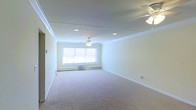 This single-floor space offers a clean, well-maintained interior with a welcoming and airy feel. The main living area is generously proportioned, featuring soft wall-to-wall carpeting that adds warmth underfoot. Crisp white crown molding runs along the perimeter of the ceiling, lending a refined architectural touch to the room. Recessed lighting is thoughtfully distributed across the ceiling, providing even illumination throughout the space, while dual ceiling fans ensure comfortable airflow year-round. Natural light filters in through a centrally positioned window, brightening the interior and creating a light, open atmosphere. Baseboard heating runs along the lower walls, offering an efficient and consistent heat source during cooler months. A window air conditioning unit provides additional climate control. The walls are finished in a soft, neutral tone that complements the light carpeting and white trim, making the space feel cohesive and move-in ready. The overall layout is open and versatile, well-suited for a variety of furnishing arrangements. The combination of classic architectural details and practical features makes this a functional and inviting single-floor space.