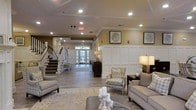 Located in Wilmington, North Carolina, this thoughtfully designed facility presents a refined and welcoming environment from the moment you enter. The main lobby serves as the heart of the space, showcasing classic architectural details including crisp white wainscoting panels, detailed crown molding, and warm neutral wall tones that create an atmosphere of understated elegance. Wide-plank hardwood-style flooring runs throughout, complementing the traditional aesthetic seamlessly. The seating area is furnished with upholstered sofas and accent chairs in neutral, textured fabrics, arranged around a central coffee table to encourage comfortable gathering and conversation. Warm table lamps add a residential quality to the lighting, supplemented by recessed ceiling lights that keep the space bright and welcoming. A staircase with a classic white balustrade anchors one side of the lobby, adding architectural interest and vertical dimension. Floor-to-ceiling glass entry doors at the far end draw in natural light, creating a bright and airy corridor. Framed botanical artwork adorns the walls, reinforcing the space's polished yet approachable character. Overall, the design balances formal architectural detailing with comfortable, livable furnishings to create a warm and functional communal environment.
