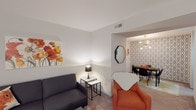 This single-floor, 2-bedroom, 2-bathroom residence features a thoughtfully designed layout that balances comfort and style. The living room serves as the central gathering space, complete with carpet flooring, a cozy seating arrangement, and a wall-mounted television setup. Vertical blinds frame a sliding door, allowing natural light to filter in. Adjacent to the living area, the dining room makes a bold statement with a striking geometric-patterned accent wall and a contemporary multi-bulb chandelier, creating an inviting atmosphere for meals and entertaining. The kitchen is visible nearby, featuring dark cabinetry and light countertops with bar seating. Both bedrooms are generously sized, offering plush carpet flooring, wide windows with horizontal blinds that let in ample natural light, and ample wall space. Each bedroom provides a calm, restful environment. The two bathrooms are finished with speckled quartz-style countertops, wide vanity mirrors, wood-look tile flooring, and tub-shower combinations. Throughout the residence, a neutral color palette is accented with warm pops of orange and modern décor elements, creating a cohesive and welcoming ambiance from room to room.