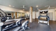 Located at 2555 Cobb Pkwy in Kennesaw, Georgia, this fitness center offers a generously sized, single-floor exercise room designed to accommodate a wide range of workout styles and fitness goals. The space features a clean, modern aesthetic defined by crisp white walls, dark rubber flooring, and a neutral gray ceiling, creating a focused and energizing atmosphere throughout.

The room is well-lit with recessed lighting and complemented by ceiling fans that help maintain air circulation during intense workouts. Wood-clad structural columns punctuate the open floor plan, adding a warm architectural contrast to the otherwise contemporary interior. Large floor-to-ceiling mirrors line one wall, visually expanding the space and supporting proper form during exercises.

Equipment is comprehensive and thoughtfully arranged. The free weights section includes a full dumbbell rack and a power rack with bench press setup. A dedicated functional training area features a multi-purpose rig stocked with battle ropes, kettlebells, medicine balls, and a punching bag. Adjustable workout benches and stability balls round out the strength-training offerings, while a row of treadmills and an upright stationary bike serve cardio needs. A wall-mounted flat-screen television adds an entertainment element. Natural light filters in through windows, enhancing the bright and inviting environment of this fully equipped facility.