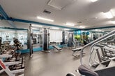 Located at 2037 Polo Gardens Dr in Wellington, FL, this fitness center offers a comprehensive workout facility within a single, efficiently organized space. The room features a durable rubber flooring surface ideal for high-traffic exercise use, with fluorescent overhead lighting ensuring a bright and well-illuminated environment throughout.

The interior is defined by a distinctive teal accent color applied along the upper wall border and architectural trim, adding a vibrant, energetic aesthetic to the space. Full-length wall mirrors line multiple sides of the room, visually expanding the area and allowing users to monitor their form during workouts.

The cardio zone is well-appointed with treadmills, elliptical trainers, and a stair climber, complemented by a wall-mounted television for entertainment. The strength training area includes a cable crossover machine, a multi-station weight machine, and a selection of selectorized equipment with adjustable benches. A dedicated free weights section features a tiered dumbbell rack stocked with a wide range of weights. Stability balls and additional accessories are also available for functional training.

Large windows along one wall allow views into an adjacent common area and permit natural light to enter, enhancing the overall atmosphere. The textured ceiling and ventilation system contribute to a comfortable training environment year-round.