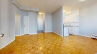 This well-appointed one-bedroom, one-bathroom unit offers a compact yet efficient layout spread across a single floor. The living room is the standout space, featuring floor-to-ceiling windows that flood the room with natural light and frame views of the surrounding urban skyline. A sliding door leads directly to a private balcony, seamlessly extending the living area outdoors. Classic parquet hardwood flooring runs throughout the main living spaces and bedroom, adding warmth and a timeless character to the interior. Soft grey walls provide a neutral backdrop throughout the unit.

The kitchen is positioned adjacent to the living area and features white cabinetry, a granite-look countertop, and white appliances, offering a clean and functional workspace. The bedroom is a well-proportioned room with its own large window overlooking the cityscape, a baseboard heating unit, and continued parquet flooring, creating a bright and comfortable retreat. A dedicated closet provides convenient storage. The hallway connects all areas of the unit efficiently and includes access to the full bathroom, which is finished with white tile surround, a bathtub, and clean white cabinetry. Overall, the unit presents a bright, airy ambiance with consistent finishes throughout.