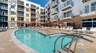 Located at 3615 Tryclan Drive in Charlotte, NC, this outdoor amenity courtyard is a standout feature of a modern multi-story residential community. The space is architecturally defined by the surrounding building's contemporary façade, which incorporates a dynamic mix of materials including brick, painted panels in neutral and yellow tones, and expansive glass-balcony railings that overlook the courtyard from multiple levels above.

At the heart of the space is a generously sized resort-style swimming pool with crystal-clear water, framed by a broad concrete pool deck. A separate spa is positioned adjacent to the main pool, offering a dedicated relaxation area. Rows of white lounge chairs are arranged along the pool's perimeter, providing ample space for sunbathing and leisure.

Beyond the pool deck, a lush turf lawn area features additional lounge seating, accented by manicured garden beds filled with flowering plants, ornamental grasses, and natural stone elements. A shaded pergola structure with seating provides a comfortable retreat from the sun.

The courtyard's enclosed design, nestled within the building's interior-facing wings, creates a sheltered and private atmosphere while still benefiting from abundant natural light and open sky views. The overall ambiance is vibrant, resort-inspired, and well-suited for outdoor relaxation and social gathering.