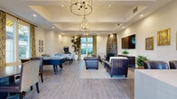 Located at 2615 E Date Palm Street in Ontario, California, this single-floor community lounge and dining space offers a well-appointed gathering area designed for both relaxation and entertainment. The interior showcases a warm, contemporary aesthetic anchored by wood-look tile flooring that flows throughout the entire space.

The living area is a standout, centered around a dramatic arched stone fireplace that rises toward the tray ceiling — a striking architectural focal point. Nearby, a wall-mounted flat-screen television and a dark wood media console complete the entertainment setup, while rich leather seating and a classic Chesterfield sofa provide comfortable places to unwind. Dual tiered chandeliers with an industrial-meets-traditional style hang above, casting warm, ambient light across the room.

The dining and game area transitions naturally from the lounge, offering a round dining table with upholstered seating alongside game tables including foosball and air hockey. Patterned drapery panels frame large windows and glass doors that open to an outdoor patio, filling the space with natural light and views of the exterior. Lush potted tropical plants are placed throughout, adding a fresh, vibrant element to the interior. Framed artwork and wall accents complete the polished, inviting atmosphere of this versatile communal space.