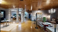 Located at 100 Foxborough Way in Richmond, Kentucky, this striking single-floor venue masterfully combines rustic charm with upscale elegance. The open-concept layout is anchored by gleaming polished concrete floors that reflect the warm ambient lighting throughout. Distinctive stacked-stone columns rise from the floor, creating natural divisions within the expansive open space while adding a timeless architectural character.

Rich wood-paneled ceilings and walls lend warmth and texture, complemented by soaring ceiling heights that allow for clerestory windows, flooding the interior with natural light. Multiple chandelier fixtures — ranging from crystal designs to modern branching styles — add a refined, decorative touch throughout.

The space is thoughtfully appointed with distinct functional areas: a welcoming reception zone featuring ornate furnishings and a curio cabinet, a lounge area with tufted leather seating and a grand stone fireplace with mounted TV and built-in shelving, and a game area centered around a full-size billiard table. A white baby grand piano and elegant seating nook add a musical and social dimension, while a well-appointed bar area rounds out the amenities. Equestrian-themed artwork and antler mounts contribute to the space's Kentucky heritage, creating an atmosphere that is both inviting and memorable.