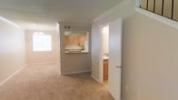 This two-floor residence features a practical and cohesive interior design with neutral tones throughout. The main living area is finished with wall-to-wall beige carpeting, creating a warm and understated atmosphere. Crown molding accents the ceilings, adding a polished architectural detail to the space.

A pass-through window connects the kitchen to the living area, offering an open and communicative layout. The kitchen is equipped with light wood cabinetry, a white refrigerator, and a countertop that extends to the pass-through, providing additional prep or casual dining space. Natural light filters into the adjoining room through a window, brightening the overall space.

Adjacent to the main living area, a doorway opens to a bathroom featuring wood-toned cabinetry, maintaining the consistent design language seen throughout the residence. The neutral wall color — a soft greige — ties the rooms together cohesively. Visible HVAC vents are integrated into the ceiling, ensuring functional climate control. The overall ambiance is clean, understated, and move-in ready.