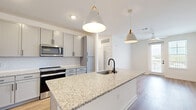 This thoughtfully designed one-bedroom, two-bathroom residence in Jacksonville, Florida offers a clean, modern aesthetic throughout its single-floor layout. The open-concept kitchen serves as the heart of the home, showcasing granite countertops, a large central island with an undermount sink and matte black faucet, gray shaker-style cabinetry, a herringbone tile backsplash, and a full suite of stainless steel appliances. Stylish pendant lighting illuminates the island, which flows seamlessly into the living area. The living space features warm hardwood-style flooring, a ceiling fan, and a glass door opening to a private balcony, allowing for natural light and outdoor views. The spacious primary bedroom is carpeted for comfort and benefits from multiple windows that invite ample natural light. A walk-in closet with wire shelving provides generous storage. Both bathrooms are finished with granite vanity tops, gray shaker cabinetry, and matte black fixtures; one includes a tub-shower combination. A second full bath offers a toilet and vanity. Additional hallways provide practical circulation throughout the unit. The consistent use of neutral tones, modern finishes, and quality materials creates a cohesive and inviting ambiance throughout this Jacksonville residence.