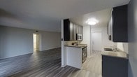 This single-floor residence offers a thoughtfully designed open-concept layout that seamlessly connects the kitchen and living area, creating a spacious and functional environment. The kitchen is a standout feature, boasting dark-toned cabinetry with a refined finish, light granite-style countertops, and stainless steel appliances including a range and over-the-range microwave. A partial wall with a breakfast bar-style counter elegantly defines the kitchen space while maintaining visual openness to the main living area. The flooring transitions effectively throughout the space — rich, dark hardwood-style planks anchor the living area, while lighter tile-style flooring is featured in the kitchen and entryway zones. The walls are finished in a clean, neutral gray tone, complementing the modern aesthetic throughout. A textured ceiling adds subtle character overhead, with flush-mount lighting providing consistent illumination. Doorways visible from the main living space lead to additional rooms, including what appears to be a bedroom and a utility or entry area, suggesting a well-connected and practical floor plan. Overall, the space presents a clean, contemporary style with a cohesive color palette and modern material choices throughout.