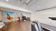 Located at 5343 Rosemead Blvd in San Gabriel, California, this fitness facility offers a thoughtfully designed workout space that maximizes its footprint with a practical and energetic layout. The main gym area features polished concrete flooring throughout, lending a clean, modern industrial aesthetic that is both durable and easy to maintain. Recessed lighting illuminates the space evenly from a textured ceiling, keeping the environment bright and functional. A striking, large-scale colorful mural adorns the exposed brick accent wall, adding a bold, artistic focal point that energizes the room. The gym is equipped with a range of cardio machines — including treadmills, ellipticals, and a stationary cycle — alongside a dedicated free weights section featuring a dumbbell rack, kettlebell rack, and an adjustable workout bench with a balance trainer. A wall-mounted air conditioning unit ensures climate control year-round. A glass-panel garage-style door on one wall allows natural light to fill the space while offering views of the surrounding area. An additional open area provides flexible space for stretching or floor exercises. The facility also includes a private, ADA-accessible bathroom finished with light wood-look flooring, clean white walls, and essential fixtures, ensuring convenience for all users.