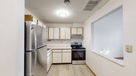 Located at 1425 N New Florissant Rd in Florissant, Missouri, this single-floor unit offers a straightforward, functional layout designed for comfortable everyday living. The kitchen is a practical, efficient space featuring light wood-trimmed white cabinetry that extends both above and below the countertops, providing ample storage. Stainless steel appliances include a full-size refrigerator and an electric range, complemented by a double stainless steel sink set into a continuous countertop. Light wood-look vinyl flooring runs throughout the kitchen, creating a clean, easy-to-maintain surface. A built-in pass-through window connects the kitchen to the adjacent living area, improving sightlines and promoting an open feel between the two spaces. The living area and bedroom are finished with neutral-toned carpet and clean white walls, contributing to a bright, understated atmosphere. The bedroom features a ceiling fan with a light fixture and a window fitted with blinds that allows natural light to filter in. Throughout the unit, white painted walls and wood-tone baseboards maintain a consistent, cohesive aesthetic. The overall layout, as seen in the dollhouse view, is compact and efficiently organized, with clearly defined zones for cooking, living, and sleeping.