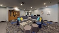 Located at 1824 E Park Pl in Milwaukee, Wisconsin, this single-floor community amenity space is thoughtfully designed to foster social interaction and relaxation. The open layout seamlessly connects a lounge area, game room, and kitchenette, all unified by a contemporary color palette of gray, blue, and yellow accents.

The lounge area is anchored by a large L-shaped sectional sofa adorned with colorful throw pillows in teal and yellow, complemented by an assortment of hexagonal ottomans and patterned poufs arranged around a central coffee table. Recessed lighting illuminates the space, while a large wall-mounted display screen serves as a focal point for entertainment and gatherings.

The game room section offers a variety of recreational options, including a pool table, shuffleboard table, and foosball table, all set atop a dynamic patterned carpet featuring blue tile accents. A dedicated workstation area with computer monitors provides a functional space for productivity within the same open environment.

The kitchenette is well-appointed with white shaker-style cabinetry reaching to the ceiling, a dark geometric tile backsplash, dark countertops, and stainless steel appliances including a microwave and mini-refrigerator. A central island with seating for multiple guests on vibrant yellow stools makes it an ideal spot for casual dining or conversation. Large-format gray tile flooring ties the kitchen aesthetic together cohesively.