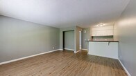 Located at 6300 N Broadway St in Kansas City, MO, this single-floor unit offers a well-organized layout comprising a living room, kitchen, and two bathrooms. The living area features warm wood-look luxury vinyl plank flooring and soft sage-green walls accented by crisp white baseboards, creating a clean and inviting atmosphere. A textured ceiling adds character throughout the space. The kitchen is positioned adjacent to the living area and includes a breakfast bar peninsula with a dark countertop, white cabinetry, and overhead lighting, providing a functional and efficient cooking space. Natural light filters in through a window with views of surrounding greenery, enhancing the open feel of the main living area. A wall-mounted HVAC unit ensures year-round climate control. The two bathrooms are finished with wood-look flooring consistent with the rest of the unit. One bathroom features a full-size soaking tub with a combination tub/shower fixture and chrome hardware, while the other includes a single-sink vanity with white cabinetry, a large mirror, and access to a spacious closet with built-in shelving and hanging rods. The neutral gray and green color palette, clean finishes, and efficient use of space make this unit a practical and comfortable residence.