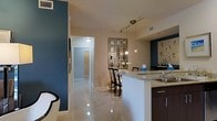 This single-floor space showcases a thoughtfully designed open-concept layout that seamlessly connects the kitchen, dining, and living areas. The kitchen features a peninsula with a granite countertop and an undermount sink, complemented by dark espresso cabinetry beneath. Pendant lighting on a gold track rail hangs above, adding a warm, contemporary touch. A decorative geometric mirror panel serves as a focal point in the dining area, where a bistro-style table and chairs create an intimate setting. The flooring throughout is a high-gloss tile that reflects light and enhances the sense of openness. Bold navy blue accent walls provide striking contrast against the neutral beige tones of the surrounding walls. A hallway leads toward additional rooms, with recessed and flush-mount ceiling lighting ensuring the space remains well-lit. Ventilation grilles are thoughtfully integrated into the ceiling. The overall ambiance balances modern aesthetics with functional design, incorporating decorative accents and quality finishes that contribute to a polished, contemporary feel throughout the space.