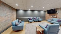 Located at 40 Vanier Dr in Guelph, Ontario, this well-appointed communal space offers a thoughtfully designed single-floor layout that blends industrial character with modern comfort. The space is defined by prominent exposed brick walls that provide a warm, textural backdrop throughout, contrasting elegantly with smooth, slate-grey painted accent walls and light hardwood-style flooring underfoot.

The layout is divided into two functional zones. The lounge area features an arrangement of contemporary light-blue upholstered sofas and armchairs accented with purple throw pillows, centered on a grey area rug. A wall-mounted flat-screen television is positioned for easy viewing from the seating. This area is ideal for casual gatherings or relaxed socializing.

The second zone serves as a meeting or dining area, anchored by a substantial rustic wood communal table surrounded by mixed metal and wood chairs. A wall-mounted display screen adds functionality for presentations or media use, while a storage cabinet provides practical utility along the wall.

Both areas share a dropped acoustic tile ceiling fitted with recessed pot lighting, ensuring consistent, even illumination throughout. A glazed partition wall and clearly marked exit doors contribute to the space's accessibility and openness. The overall ambiance is inviting and functional, making this space well-suited for community gatherings, meetings, or collaborative activities.