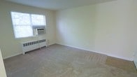 This single-floor space presents a straightforward and functional layout, highlighted by a generously sized main room with neutral, cream-colored walls that create a clean and understated atmosphere. The room is finished with wall-to-wall carpeting in a light beige tone, lending a sense of warmth and comfort underfoot. A window fitted with a through-the-wall air conditioning unit provides both natural light and climate control, while a baseboard radiator beneath the window ensures efficient heating during cooler months. White baseboards run along the perimeter of the room, adding a modest architectural trim detail that complements the overall minimal aesthetic. The walls are smooth and well-maintained, offering a blank canvas for a variety of furnishing styles. The space benefits from a calm, quiet ambiance, with the soft natural light filtering through the window contributing to an open and airy feel. The simplicity of the design ensures versatility, making the room adaptable for use as a living area, bedroom, or home office. Overall, the space is practical, well-appointed with essential climate systems, and presents a neutral foundation suitable for a range of occupants and uses.