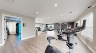 Located at 601 SW Sapphire Dr in Bentonville, Arkansas, the Touchstone Village fitness center is a thoughtfully designed, single-floor community gym that provides residents with a comprehensive workout environment. The space features light wood-look flooring throughout, recessed lighting, and a neutral color palette of warm gray and deep charcoal accent walls — including a bold Touchstone Village logo wall that anchors the strength training area.

The main workout floor is generously sized, accommodating a wide array of equipment including treadmills, upright and recumbent stationary bikes, an elliptical, a multi-function cable machine, a weight rack with dumbbells, and a flat bench. Motivational wall lettering — "The Only Bad Workout Is The One That Didn't Happen" — adds an energizing atmosphere to the space.

Multiple windows fitted with white blinds line the walls, allowing ample natural light to fill the room. A set of glass doors opens directly to an outdoor area, offering views of what appears to be a community pool and green space beyond.

A separate entry foyer area includes a small refreshment station with bar stools, and a dedicated tanning room — marked with clear signage — is accessible through a private door. The overall ambiance is clean, modern, and well-maintained, creating a motivating environment for all fitness levels.