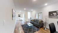 This stylish single-floor residence offers a seamlessly connected layout designed for comfortable, modern living. The open-plan living area serves as the heart of the space, anchored by warm hardwood flooring that extends throughout and creates a sense of continuity. Crisp white walls and recessed lighting combine to produce a bright, airy atmosphere, while large windows invite an abundance of natural light.

The living room is thoughtfully appointed, featuring a plush charcoal sofa paired with a geometric patterned area rug and a sleek glass coffee table. Two tall decorative mirrors with silver frames add depth and elegance to the space, reflecting light and enhancing the room's open feel. A set of white accent chairs provides additional seating, ideal for entertaining or relaxing.

The corridor leading deeper into the residence continues the home's clean aesthetic, hinting at well-proportioned additional rooms beyond. Neutral tones, recessed ceiling lights, and consistent flooring tie the entire space together cohesively.

Overall, the space strikes a balance between refined minimalism and functional comfort, making it an inviting environment with a polished, contemporary character throughout.