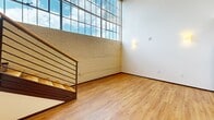 This two-story loft at 675 N Main St in downtown Winston-Salem blends industrial character with modern finishes. The main level features warm hardwood flooring, exposed concrete ceilings with track lighting, a structural column, and large industrial-style windows that flood the space with natural light. The kitchen is equipped with rich dark wood cabinetry, granite countertops, a mosaic tile backsplash, and stainless steel appliances. A stylish bathroom showcases dual vessel sinks on a granite vanity with slate tile flooring. The upper level hosts the private bedroom with a closet, an additional bathroom, and a dedicated laundry area, all connected by a hardwood staircase framed by an exposed brick accent wall.
