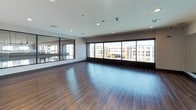 This single-floor yoga and fitness studio is thoughtfully designed to support a variety of movement practices in a clean, modern environment. The main studio space is expansive and open, featuring rich hardwood flooring that extends throughout and provides an ideal surface for yoga, dance, and other fitness activities. A full wall of floor-to-ceiling mirrors lines one side of the studio, allowing practitioners to monitor their form and creating a sense of added depth and openness. Opposite the mirrors, large picture windows span the length of the room, flooding the space with abundant natural light and offering views of the surrounding urban streetscape. Recessed ceiling lighting supplements the natural light for evening or low-light sessions. A large flat-screen television mounted above the mirror wall provides visual guidance for instructors and participants alike, with a tripod stand available for additional equipment setup. The welcoming entry area features warm peach-toned walls adorned with motivational wall art reading "FREE" and "Go with the Flow," setting an encouraging tone for visitors. Rolled yoga mats are neatly stored in the entry alongside open shelving, ensuring easy access to equipment. The studio's overall ambiance is energizing yet serene, combining contemporary finishes with an airy, light-filled atmosphere.