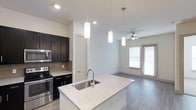 This single-floor space showcases a thoughtfully designed open-concept layout that seamlessly connects the kitchen and living area. The kitchen is a standout feature, anchored by a spacious island with a striking granite countertop, an undermount sink, and a sleek modern faucet. Pendant lighting hangs overhead, adding a warm, focused glow to the workspace. Dark espresso cabinetry provides bold contrast against the light walls, while stainless steel appliances — including a range and built-in microwave — complete the contemporary aesthetic. A tasteful tile backsplash adds texture and visual interest behind the range. The flooring transitions smoothly from the kitchen into the living area, where wood-look planks lend warmth and continuity throughout the space. The living area benefits from natural light streaming through a large window and a glass door, creating a bright and inviting atmosphere. A ceiling fan with a light fixture keeps the space comfortable year-round. Clean white walls and neutral tones throughout create a calm, versatile backdrop that suits a variety of interior styles. The overall ambiance is modern, open, and functional — ideal for both everyday living and entertaining.