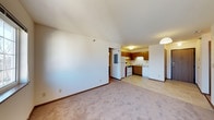 This single-floor space features an open-concept layout where the living area connects seamlessly to the kitchen, creating a functional and cohesive environment. The living area is finished with wall-to-wall beige carpeting that adds a sense of warmth, while the smooth, textured ceiling and neutral white walls provide a clean, understated backdrop. A pendant light fixture marks the transition into the kitchen area, offering ambient illumination.

The kitchen is efficiently designed with warm wood-toned cabinetry that lines both the upper and lower sections of the space. White appliances, including a refrigerator and a range with an overhead microwave or vent hood, are neatly integrated into the layout. The kitchen floor transitions to a light-toned tile, offering a practical surface in the cooking and food preparation zone. A double sink sits beneath the cabinetry, contributing to the kitchen's utilitarian design.

A hallway visible from the living area leads to additional rooms, suggesting a clear separation of living and private spaces. The overall ambiance is modest and functional, with a straightforward design suited for everyday living.