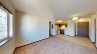 This single-floor space features an open-concept layout where the living area connects seamlessly to the kitchen, creating a functional and cohesive environment. The living area is finished with wall-to-wall beige carpeting that adds a sense of warmth, while the smooth, textured ceiling and neutral white walls provide a clean, understated backdrop. A pendant light fixture marks the transition into the kitchen area, offering ambient illumination.

The kitchen is efficiently designed with warm wood-toned cabinetry that lines both the upper and lower sections of the space. White appliances, including a refrigerator and a range with an overhead microwave or vent hood, are neatly integrated into the layout. The kitchen floor transitions to a light-toned tile, offering a practical surface in the cooking and food preparation zone. A double sink sits beneath the cabinetry, contributing to the kitchen's utilitarian design.

A hallway visible from the living area leads to additional rooms, suggesting a clear separation of living and private spaces. The overall ambiance is modest and functional, with a straightforward design suited for everyday living.