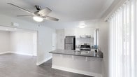 This single-floor residence offers a thoughtfully designed open-concept layout that seamlessly connects the kitchen and living areas, creating a spacious and inviting atmosphere. The kitchen is a standout feature, showcasing crisp white cabinetry, dark granite-style countertops, and a full suite of stainless steel appliances including a refrigerator, gas range with oven, and a built-in microwave. A peninsula breakfast bar extends from the kitchen, providing additional counter space and casual seating — ideal for both dining and entertaining. The kitchen is well-lit with recessed ceiling lighting that enhances the clean, modern aesthetic. Rich dark hardwood-style flooring runs throughout the space, offering a warm contrast to the light, neutral-toned walls. The living area is equipped with a ceiling fan with integrated lighting, adding both comfort and functionality. A doorway visible from the main living space leads to additional rooms, suggesting a practical and well-organized floor plan. Natural light filters in through a window near the cooking area, brightening the space. Overall, the unit presents a clean, contemporary feel with quality finishes throughout.