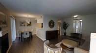 This single-story residence offers an open-concept layout that connects the living, dining, and kitchen areas into one cohesive and functional space. The interior is finished with rich dark hardwood-style flooring that runs consistently throughout, creating a warm and grounded aesthetic against the soft gray walls.

The kitchen is positioned behind a raised white breakfast bar that provides a natural partition while maintaining visual openness to the rest of the space. A stainless steel refrigerator and dark cabinetry are visible, adding contrast to the light countertops and white upper cabinetry.

Adjacent to the kitchen, the dining area is defined by a sleek dining table with white chairs and a modern pendant light overhead, accompanied by abstract wall art that adds a contemporary touch. Across the space, the living area features a textured area rug, woven accent chairs, and a round ottoman, creating a relaxed seating arrangement.

A decorative sunburst mirror mounted on the central wall serves as a focal point, while a tall potted palm adds a natural, organic element. The entry door and hallway leading to additional rooms are visible, suggesting a well-organized and practical floor plan throughout.