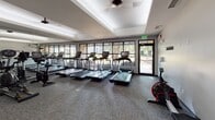 Located at 201 Carolina Point Pkwy in Greenville, South Carolina, this fitness center offers a well-appointed, single-floor workout facility designed to support a well-rounded exercise routine. The space is finished with durable carpet flooring throughout and features a clean, modern aesthetic with neutral gray walls and a tray ceiling detail that adds architectural depth.

The cardio section is a standout feature, housing a row of Life Fitness treadmills lined along a wall of large windows that frame views of the surrounding greenery and outdoor environment. Full-length mirrors run along an adjacent wall, enhancing the sense of openness and allowing users to monitor their form. Complementing the treadmills is an assortment of stationary bikes and elliptical trainers, providing varied cardio options.

A dedicated strength training area includes a well-stocked dumbbell rack with a full range of free weights alongside a weight bench, offering essential resistance training equipment. Additional cardio machines, including rowing-style bikes, are distributed throughout the space.

Natural light filters generously through the large window panels, creating a bright and energizing atmosphere. Glass-panel dividers provide visual connection between zones while maintaining an open feel. The facility is well-equipped with safety features including clearly marked emergency exits, making it a practical and comfortable space for fitness enthusiasts.