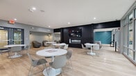 Located at 1450 E Summit St in Kent, Ohio, this contemporary community amenity space spans a generous single floor, thoughtfully designed to accommodate a variety of social and professional activities. Light wood-style flooring flows throughout, complementing the modern, neutral color palette accented by bold teal and deep navy design elements.

The main gathering area features multiple round white pedestal tables with stylish grey chairs, ideal for casual dining or socializing. Floor-to-ceiling glass walls and large sliding doors flood the space with abundant natural light while offering direct views of a covered outdoor patio and surrounding tree-lined grounds. A dramatic black accent wall with a linear fireplace and mounted flat-screen TV serves as a striking focal point, with adjacent cozy seating alcoves for quieter moments.

A dedicated co-working room stands out with vibrant teal walls, a live-edge communal table, yellow and brown task chairs, and dual wall-mounted displays — a well-equipped space for collaboration. A large sectional sofa area provides a relaxed lounge environment anchored by a geometric teal feature wall.

Completing the space is a café-style coffee bar with white cabinetry, a beverage cooler, and a coffee station, making this a fully equipped, inviting hub for community life.