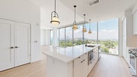 This contemporary high-rise unit is defined by its clean architectural lines, bright white palette, and an abundance of natural light streaming through expansive floor-to-ceiling glass walls. The open living area flows seamlessly toward sweeping panoramic views of a city skyline and tree-lined urban landscape below, creating a striking visual connection to the outdoors.

The kitchen is a central highlight, featuring a generously proportioned white quartz island with an integrated sink, flat-front cabinetry extending to the ceiling, a subway tile backsplash, stainless steel appliances, and a series of modern arched pendant lights overhead. An in-unit laundry area is conveniently tucked nearby.

Bedroom spaces are bright and airy, with light wood flooring and direct access to balcony areas that continue the outdoor views. The primary bedroom is accompanied by a spacious walk-in closet outfitted with wire shelving systems along multiple walls.

The primary bathroom is particularly refined, offering a freestanding soaking tub, dual vanities with white cabinetry, globe wall sconces, and large-format grey tile flooring. A separate water closet provides added privacy. A utility area with open shelving and an electrical panel rounds out the functional elements of this well-appointed space.