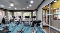 This single-floor fitness center is designed to provide a functional and inviting workout environment. The space is outfitted with a selection of Life Fitness cardio equipment, including treadmills, elliptical trainers, and a recumbent bike, catering to a range of exercise preferences and fitness levels. A standout feature of the room is its wraparound bank of large windows, which flood the space with abundant natural light and offer views of the surrounding outdoor greenery and pool area, creating an energizing atmosphere for workouts. The recessed ceiling lighting complements the natural light, ensuring the space remains bright and well-lit at all times. Underfoot, a bold geometric-patterned carpet in shades of teal and blue adds a vibrant, modern aesthetic that distinguishes the space visually. The layout is thoughtfully arranged to allow comfortable movement between equipment, while the neutral grey walls and trim provide a clean, contemporary backdrop. A glass partition on one side offers a visual connection to an adjacent area, enhancing the open feel of the space. Overall, the fitness center strikes a balance between practicality and design, making it an appealing destination for daily exercise.
