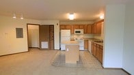 This single-floor space is designed with a practical, open-concept layout that integrates the kitchen and living area into one cohesive space. The kitchen is outfitted with warm honey-toned wood cabinetry that provides ample upper and lower storage, complemented by white appliances including a refrigerator, range, built-in microwave, and dishwasher. A freestanding central island adds additional workspace and serves as a natural divider between the kitchen and the adjoining living area. The kitchen flooring consists of light neutral tile, which transitions into wall-to-wall carpeting in the living space, offering a comfortable and casual feel underfoot. Flush-mount ceiling lighting illuminates both areas effectively. Adjacent to the main living space, a doorway leads to what appears to be a closet with shelving or hanging storage, offering useful organizational capacity. The entry door is also visible from this area, with a floor-level vent indicating a forced-air climate system. The overall color palette is neutral and understated, with beige and warm wood tones throughout, creating a functional and move-in-ready environment.