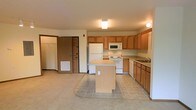 This single-floor space is designed with a practical, open-concept layout that integrates the kitchen and living area into one cohesive space. The kitchen is outfitted with warm honey-toned wood cabinetry that provides ample upper and lower storage, complemented by white appliances including a refrigerator, range, built-in microwave, and dishwasher. A freestanding central island adds additional workspace and serves as a natural divider between the kitchen and the adjoining living area. The kitchen flooring consists of light neutral tile, which transitions into wall-to-wall carpeting in the living space, offering a comfortable and casual feel underfoot. Flush-mount ceiling lighting illuminates both areas effectively. Adjacent to the main living space, a doorway leads to what appears to be a closet with shelving or hanging storage, offering useful organizational capacity. The entry door is also visible from this area, with a floor-level vent indicating a forced-air climate system. The overall color palette is neutral and understated, with beige and warm wood tones throughout, creating a functional and move-in-ready environment.