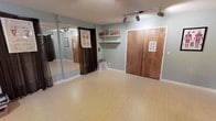 This single-floor studio space is thoughtfully designed to support movement-based activities such as dance, yoga, or personal fitness training. The open floor plan is finished with smooth, light-toned hardwood-style flooring that provides an ideal surface for a wide range of physical activities.

A full-length mirrored wall along one side of the room is a defining architectural feature, allowing practitioners to monitor form and technique during sessions. The mirrors are framed neatly and span a generous portion of the wall, maximizing their functional utility. Adjacent to the mirrors, a dark curtain offers the option to section off or cover a private changing or storage area.

The walls are painted in a soft, muted blue-gray tone that contributes to a calm and focused atmosphere. Track lighting mounted on the ceiling provides adjustable, directional illumination throughout the space, ensuring consistent brightness during use. Built-in wall shelving offers convenient storage for supplies and personal items.

A set of solid wood double doors serves as the main entry point, lending the space a sturdy, welcoming presence. Informational posters are displayed near the entrance, adding an educational touch appropriate for a wellness or instruction-oriented environment. Overall, the studio balances practicality with a clean, inviting aesthetic suited for professional or personal use.