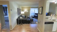 This single-floor space offers a thoughtfully designed interior that balances modern aesthetics with everyday comfort. The living area serves as the centerpiece, anchored by warm hardwood flooring that extends throughout the space and adds a natural, inviting tone to the overall design. A large window at the far end of the room floods the area with natural light, framing a pleasant outdoor view and enhancing the sense of openness. The lounge area is arranged with a deep charcoal sectional sofa complemented by a tufted ottoman and a coordinating armchair, creating a comfortable and cohesive seating arrangement. A geometric patterned area rug defines the space, while a round glass side table and a woven pouf add functional yet decorative accents. Warm ambient lighting is provided by a pair of floor lamps, contributing to the cozy atmosphere. The walls are decorated with framed artwork, adding a personal and curated touch to the space. A narrow entryway with clean lines and neutral tones leads into the living area, reinforcing the space's streamlined and contemporary feel. Overall, the design emphasizes a clean, modern style with warm undertones and practical furnishings.
